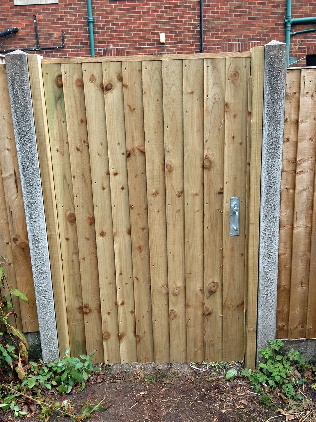 Closeboard timber garden gate between concrete posts, with a brick wall behind